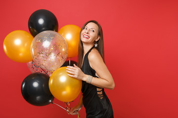 Fototapeta na wymiar Joyful young woman in little black dress celebrating holding air balloons isolated on red background. St. Valentine's, International Women's Day, Happy New Year, birthday mockup holiday party concept.