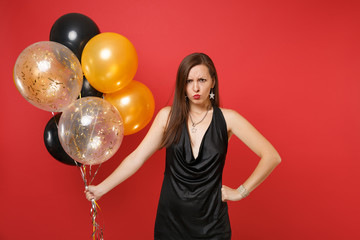 Dissatisfied offended woman in little black dress holding air balloons while celebrating isolated on red background. International Women's Day, Happy New Year, birthday mockup holiday party concept.