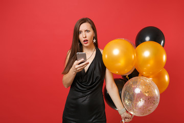Puzzled young woman in little black dress holding air balloons, using mobile phone while celebrating isolated on red background. Valentine's Day, Happy New Year, birthday mockup holiday party concept.