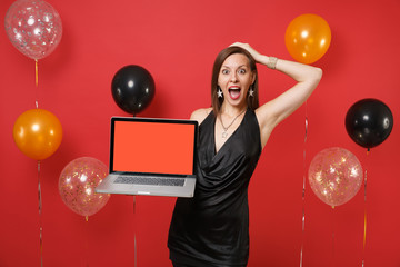 Excited girl in black dress putting hand on head holding laptop pc computer with blank black empty screen on bright red background air balloons. Happy New Year, birthday mockup holiday party concept.