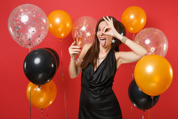 Funny young girl in black dress celebrating holding glass of champagne, showing tongue, OK sign near eyes on bright red background air balloons. Happy New Year, birthday mockup holiday party concept.