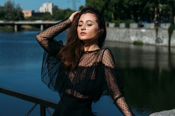 beautiful girl, brunette, in the summer by the river in the city, in a black dress. standing alone by the river, the sun shining in the face, closed her eyes, rest and tourism.