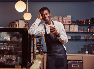 African barista smiling at camera relaxing after workday with coffee while leaning on the counter at coffee shop.