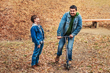 Father laughs his son for a walk in the autumn park. Dad rides his son's scooter