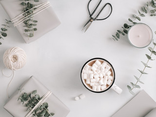 A mug with hot chocolate and marshmallow among gift boxes and eucalyptus on a white table. The concept of wintertime and wrapping gifts. Flat lay; top view; minimalist style.