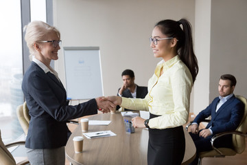 Smiling middle aged businesswoman handshake Asian employee congratulating with promotion, happy female boss shake hand of millennial woman worker greeting with good work results. Reward concept