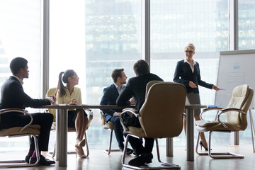 Positive middle-aged female coach or mentor training employees, making flipchart presentation at meeting, smiling mature businesswoman present project or idea to colleagues at office negotiations