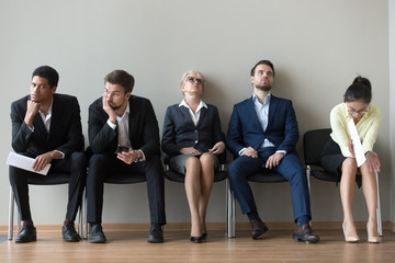 Multiethnic job candidates in queue tired of long waiting in office corridor, diverse work applicants sit on chairs feel exhausted expecting their turn for interview. Employment, hiring, HR concept
