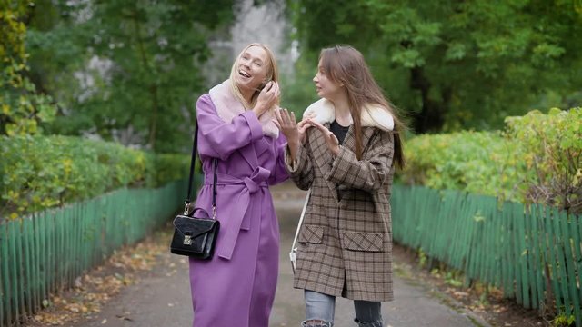 two cheerful women friends are wearing warm coat are walking in autumn day on alley in park and chatting