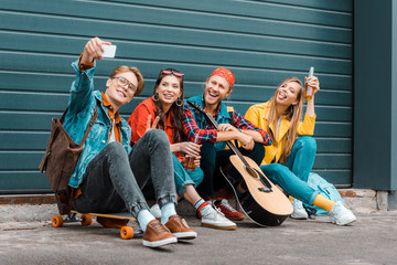 young friends with beer, skateboard and guitar taking selfie on smartphone