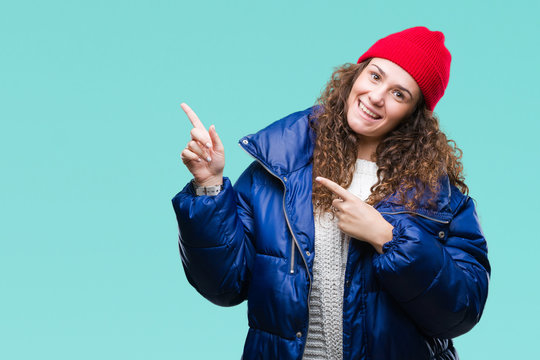 Beautiful young brunette curly hair girl wearing winter coat, wool cap and sweater over isolated background smiling and looking at the camera pointing with two hands and fingers to the side.