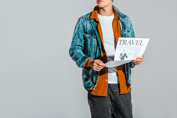 cropped view of hipster in denim jacket reading travel newspaper isolated on grey