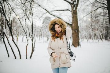 A beautiful young Caucasian woman stands in a snowy park in a jacket with a hood and fur on her head in jeans and holds a pair of white skates on her shoulder