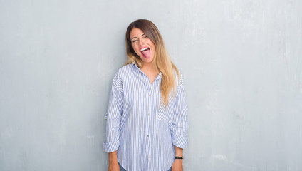 Young adult woman over grey grunge wall wearing fashion business outfit sticking tongue out happy with funny expression. Emotion concept.