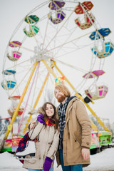 Fototapeta premium Young couple in love with a happy young man with a beard and a woman on a background of a fucking colossus, a Ferris wheel resting, a date in an amusement park in the winter in the Christmas holidays