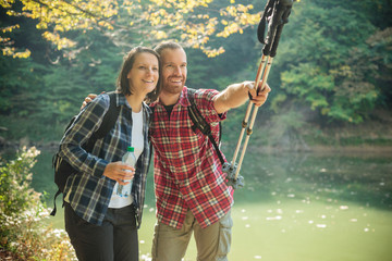 Smiling happy young couple in checkered shirts hiking along the lake shore, embracing each other. Man is pointing with his hand. Forest on a beautiful autumn day. Healthy and active outdoor lifestyle
