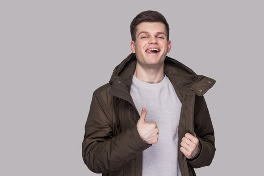 Portrait of young funny handsome man standing in light gray shirt and dark green parka looking at camera and showing thumbs up.. indoor studio shot, isolated on gray background.