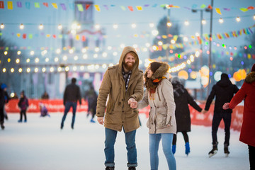 Young couple in love Caucasian man with blond hair with long hair and beard and beautiful woman have fun, active date skating on ice scene in town square in winter on Christmas Eve