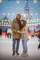 Young couple in love Caucasian man with blond hair with long hair and beard and beautiful woman have fun, active date skating on ice scene in town square in winter on Christmas Eve