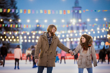 Young couple in love Caucasian man with blond hair with long hair and beard and beautiful woman have fun, active date skating on ice scene in town square in winter on Christmas Eve