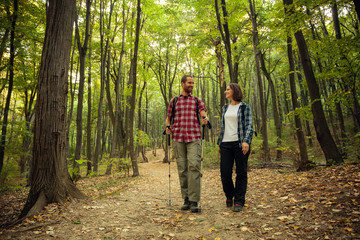 Obraz premium Loving couple wearing checkered shirts hiking side by side through forest on a beautiful autumn day, looking at each other. Healthy and active outdoor lifestyle