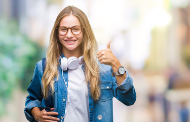 Young beautiful blonde student woman wearing headphones and glasses over isolated background doing happy thumbs up gesture with hand. Approving expression looking at the camera with showing success.