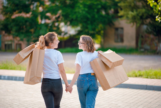 Rear view of young girls in white shirts and jeans with shopping bags walking on the street holding hands. Two female friends