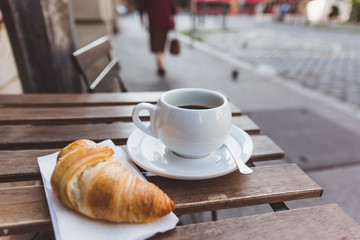 Breakfast with black coffee and croissants on the wooden table in an outdoor cafe. City on a background