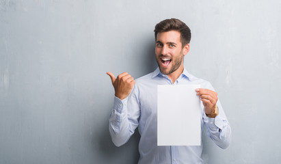 Handsome young man over grey grunge wall holding blank paper sheet contract pointing and showing with thumb up to the side with happy face smiling