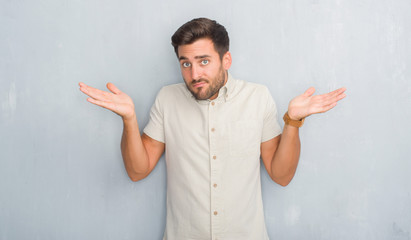 Handsome young man over grey grunge wall wearing summer shirt clueless and confused expression with arms and hands raised. Doubt concept.