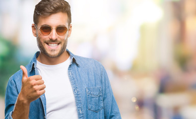 Obraz premium Young handsome man wearing sunglasses over isolated background doing happy thumbs up gesture with hand. Approving expression looking at the camera with showing success.