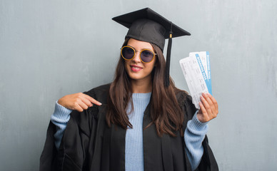 Young brunette woman over grunge grey wall wearing graduate uniform holding boarding pass with...