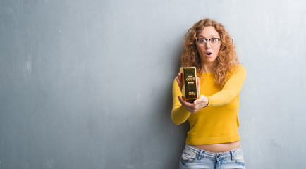 Young redhead woman over grey grunge wall holding gold ingot scared in shock with a surprise face,...
