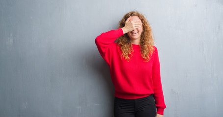 Young redhead woman over grey grunge wall wearing red sweater smiling and laughing with hand on face covering eyes for surprise. Blind concept.