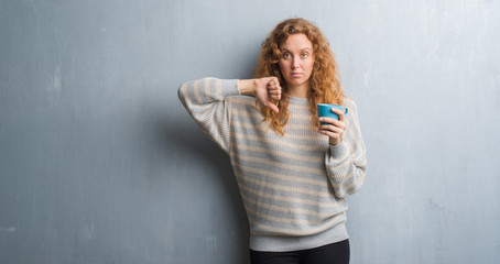 Fototapeta premium Young redhead woman over grey grunge wall drinking a cup of coffee with angry face, negative sign showing dislike with thumbs down, rejection concept