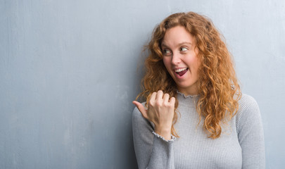 Young redhead woman over grey grunge wall pointing and showing with thumb up to the side with happy face smiling