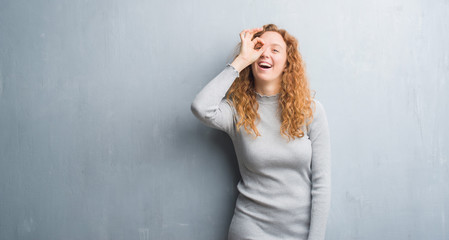 Young redhead woman over grey grunge wall with happy face smiling doing ok sign with hand on eye looking through fingers
