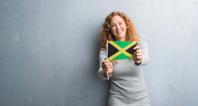 Young redhead woman over grey grunge wall holding flag of Jamaica with a happy face standing and smiling with a confident smile showing teeth