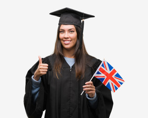 Young hispanic woman wearing graduated uniform holding flag of united kingdom happy with big smile doing ok sign, thumb up with fingers, excellent sign