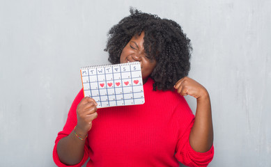 Young african american woman over grey grunge wall holding menstruation calendar screaming proud...