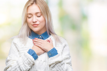 Young blonde woman wearing winter sweater over isolated background smiling with hands on chest with closed eyes and grateful gesture on face. Health concept.