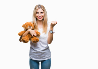 Young beautiful blonde woman holding teddy bear over isolated background screaming proud and celebrating victory and success very excited, cheering emotion