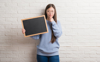 Young Chinese woman over brick wall holding blackboard serious face thinking about question, very confused idea