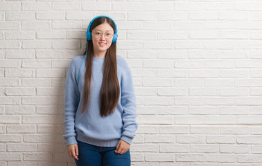 Young Chinese woman over brick wall wearing headphones with a happy face standing and smiling with a confident smile showing teeth