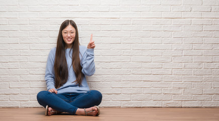 Young Chinese woman sitting on the floor over brick wall showing and pointing up with finger number one while smiling confident and happy.