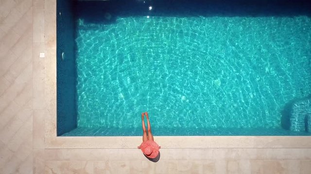 Aerial view of young girl in sunhat sitting by swimming pool.