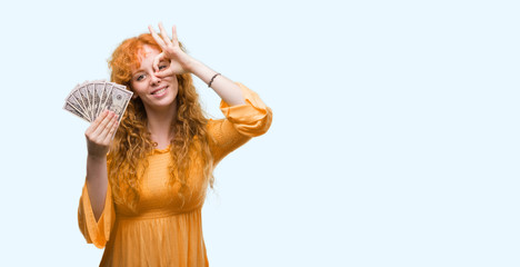 Young redhead woman holding dollars with happy face smiling doing ok sign with hand on eye looking through fingers