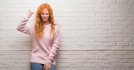 Young redhead woman standing over brick wall angry and mad raising fist frustrated and furious while shouting with anger. Rage and aggressive concept.