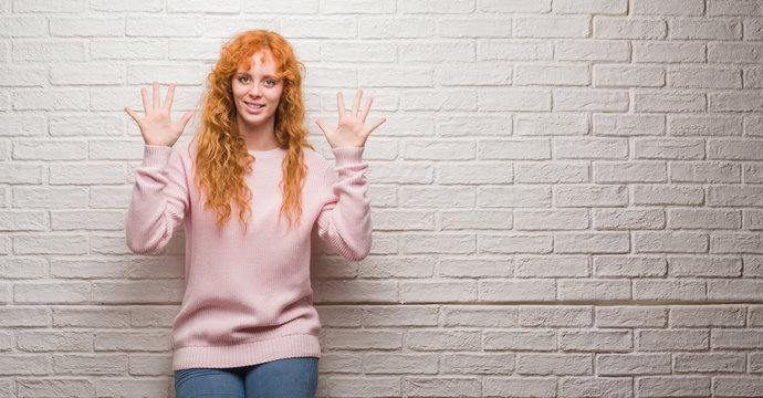 Young redhead woman standing over brick wall showing and pointing up with fingers number ten while smiling confident and happy.