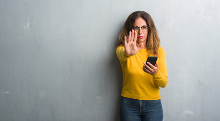 Middle age hispanic woman using smartphone with open hand doing stop sign with serious and...